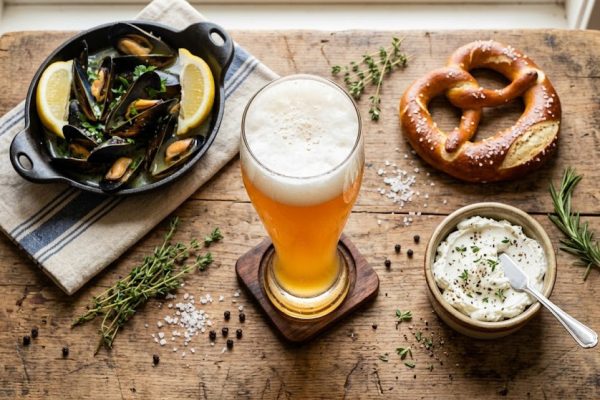 Hazy wheat beer in a weizen glass paired with mussels, a soft pretzel, and goat cheese on a wooden table, illustrating ideal wheat beer food pairings