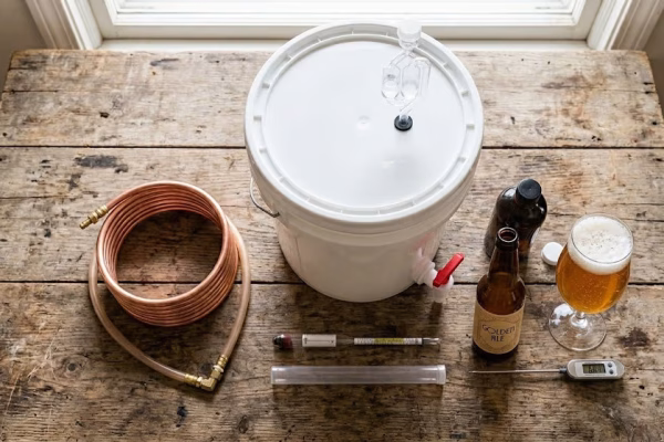 Beginner homebrewing equipment laid out on a wooden table including a fermenter bucket, airlock, auto-siphon, hydrometer, and Star San sanitizer