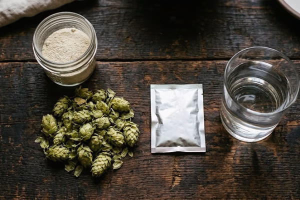 The four ingredients needed to brew beer at home arranged on a wooden surface including a jar of dry malt extract, fresh hop cones, a packet of dry yeast, and a glass of clear water
