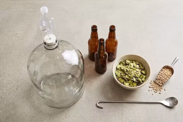 Homebrew beer making kit flat lay with glass fermenter, brown bottles, dried hops, and grain on a kitchen counter — Father's Day gift for homebrewing dads