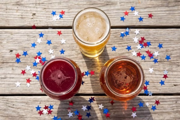 Three craft beers in red, white, and blue glasses arranged on a wooden picnic table with Fourth of July decorations